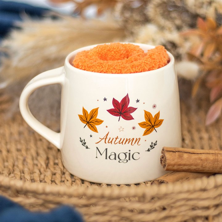 White mug with 'Autumn Magic' text and leaf designs, filled with orange fabric, on a woven surface.
