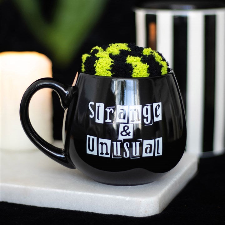 Black mug with 'Strange & Unusual' text and a knitted item inside, on a blurred background.
