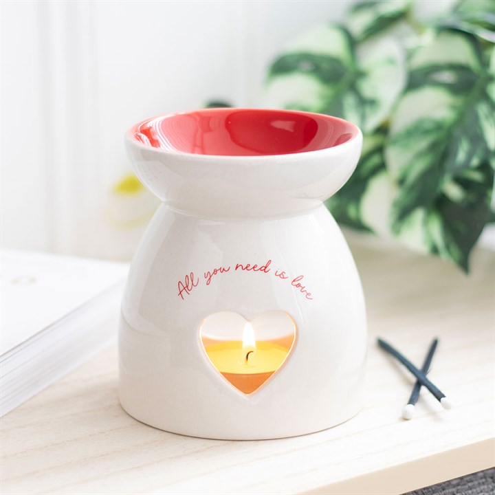 White ceramic wax warmer oil burner diffuser with red top and 'All you need is love' text, placed on a surface with a blurred plant in the background.