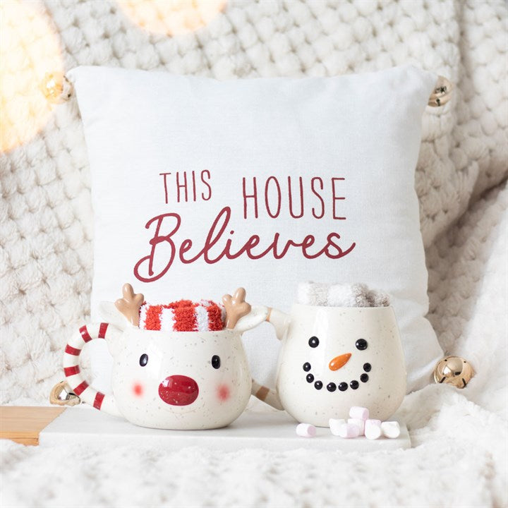 Decorative pillow with 'This House Believes' text, surrounded by a reindeer mug and snowman ornament on a textured surface.