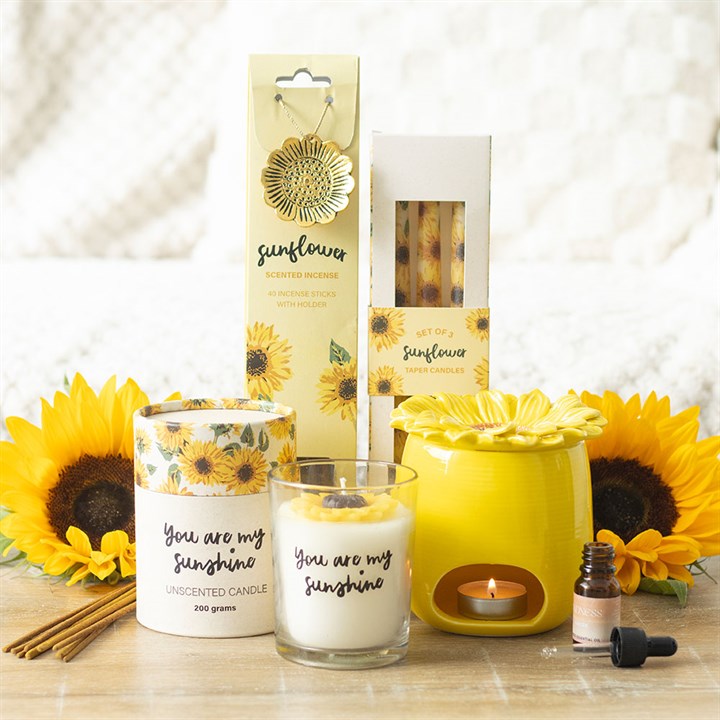Sunflower-themed candles, diffuser, and essential oil on a wooden surface with sunflowers in the background.