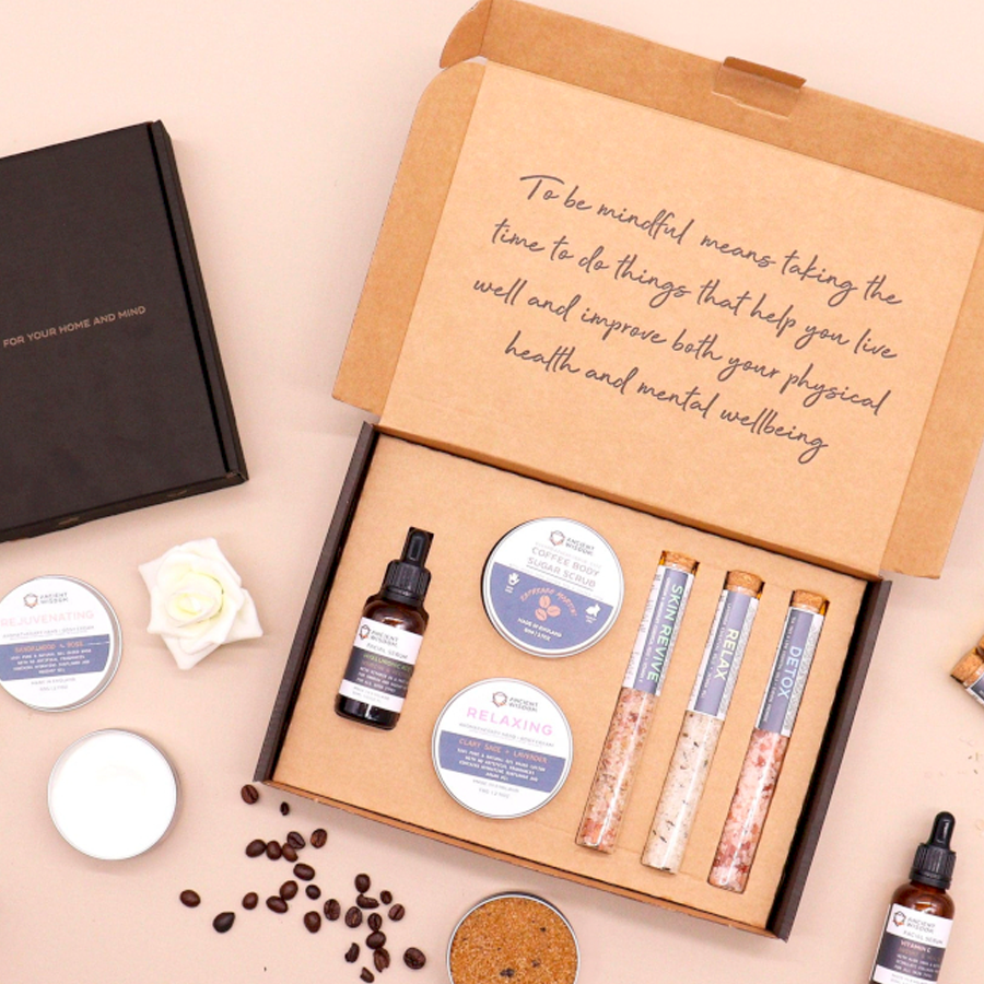 A gift box open to reveal various self-care products including a coffee body sugar scrub, a lavender hand and body cream, bath salt vials, and a hyaluronic acid facial serum. The box is set against a neutral background with scattered plant essential oil bottles and ingredients.
