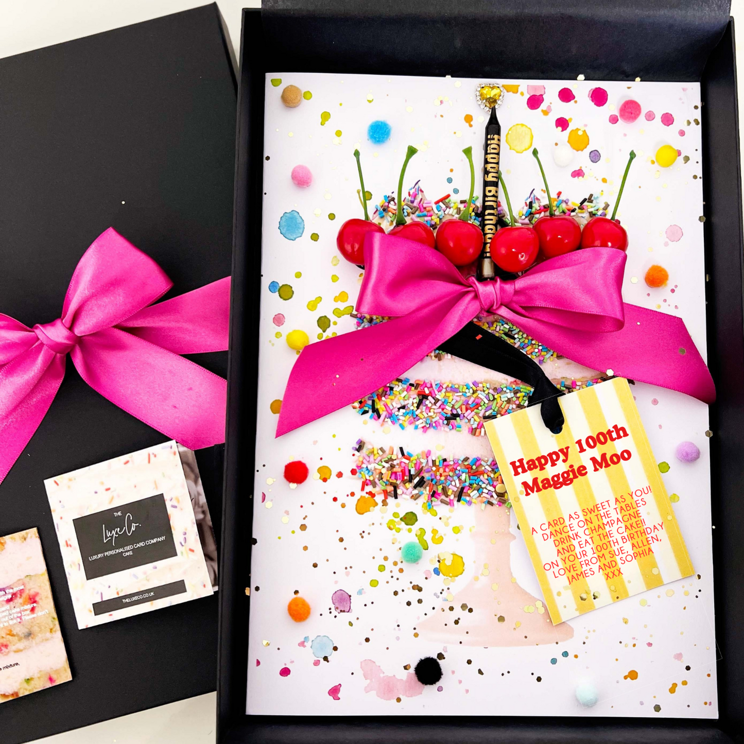 Gift box with a cake-themed 100th birthday card, pink bow, and colorful confetti on a white background.