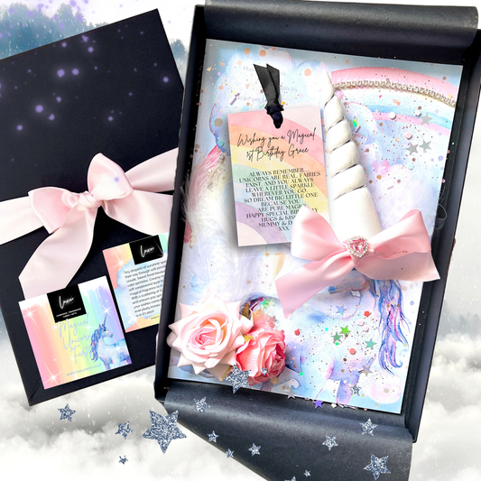 A magical Unicorn first birthday card and unicorn-themed birthday gift set displayed in a black box, featuring a pastel unicorn illustration, pink bows, rose decorations, and a small card. A matching black box with a pink bow is shown beside it on a soft cloud background.
