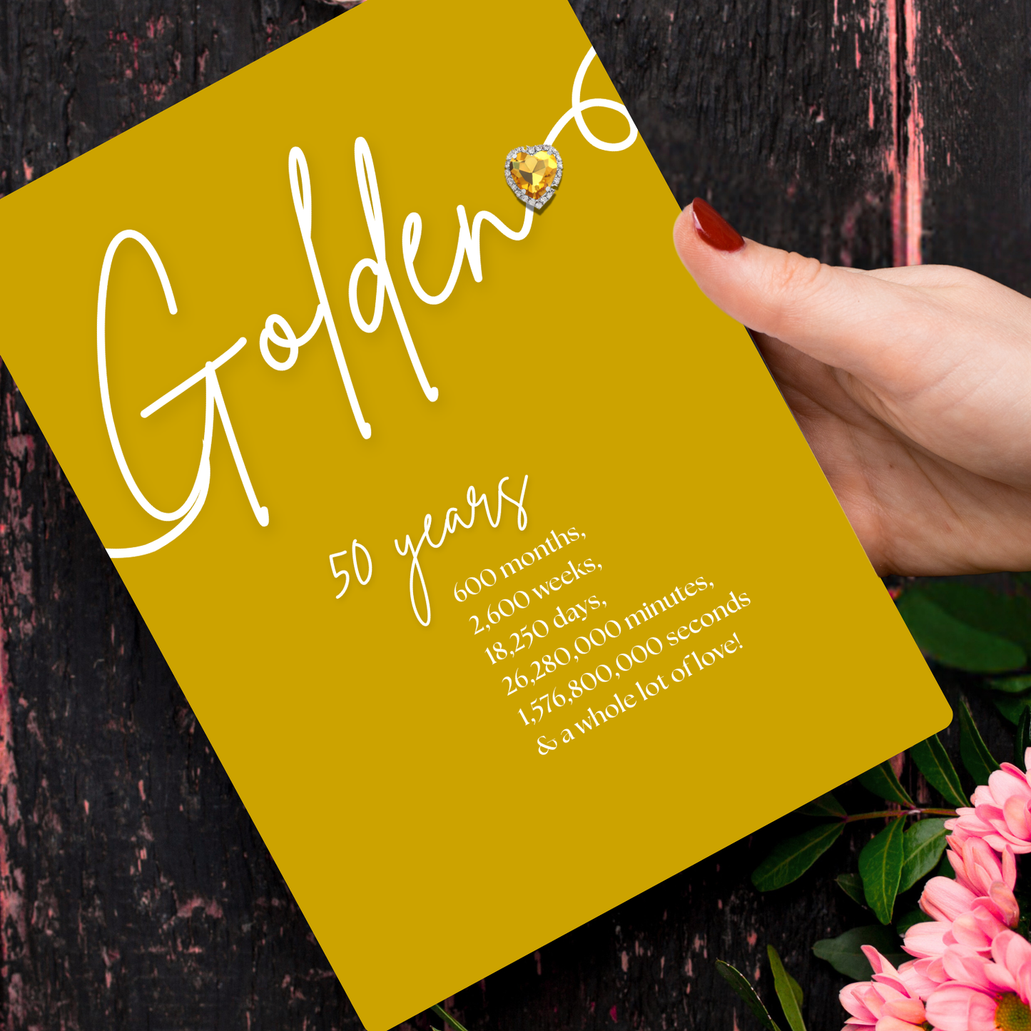 Personalised A5 Swirly Sparkly Diamante Golden Heart Anniversary Card with Yellow card with 'Golden 50 years' text, held by a hand with red nail polish, against a dark background with pink flowers.
