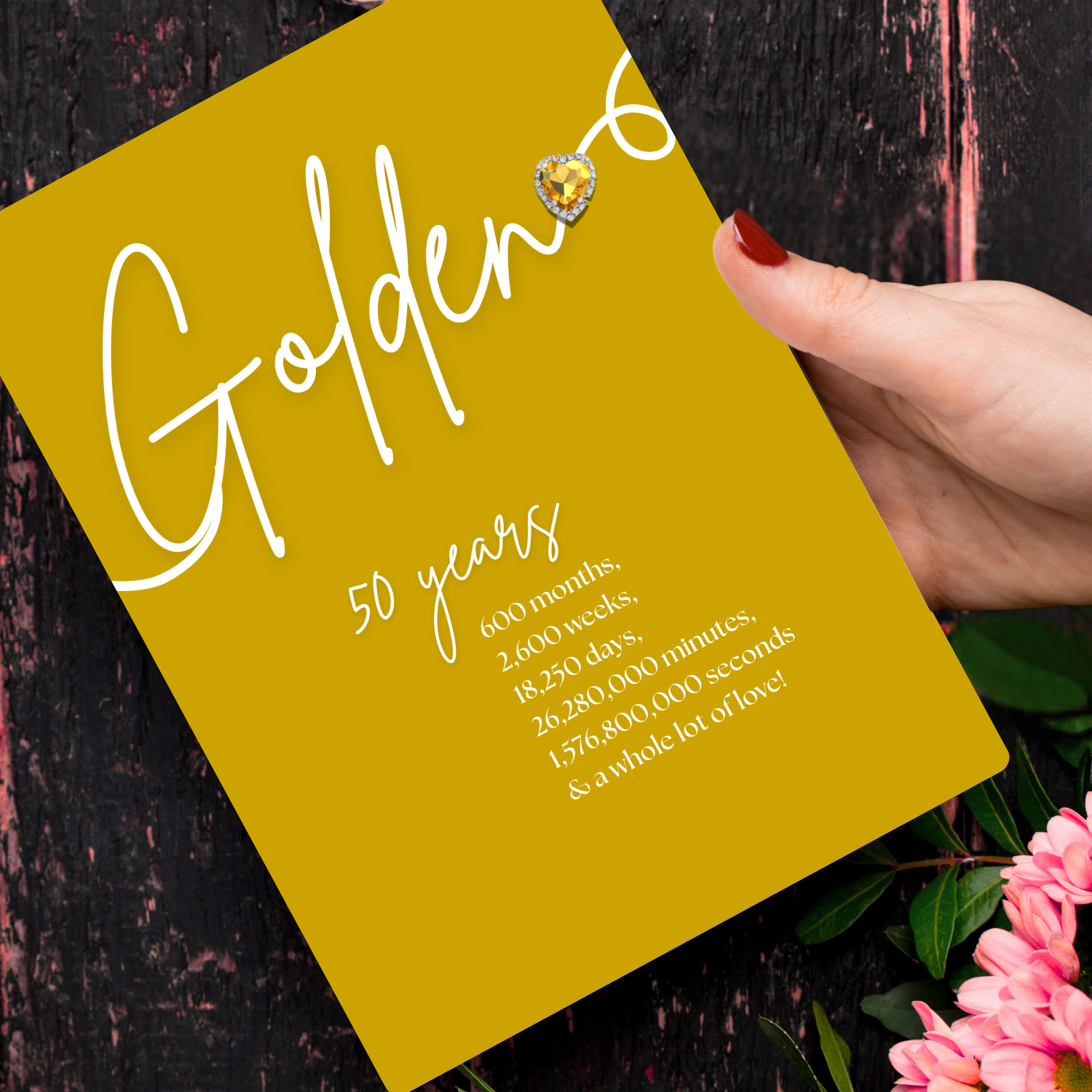 Personalised A5 Swirly Sparkly Diamante Golden Heart Anniversary Card with Yellow card with 'Golden 50 years' text, held by a hand with red nail polish, against a dark background with pink flowers.