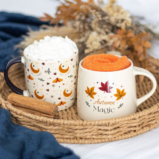 Two mugs with autumn-themed designs on a woven tray with whipped cream and an orange cloth.