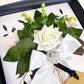A handmade wedding anniversary card featuring a white rose, green leaves, and a sparkly crystal on a silky satin ribbon sash, presented in a black and white photo frame.