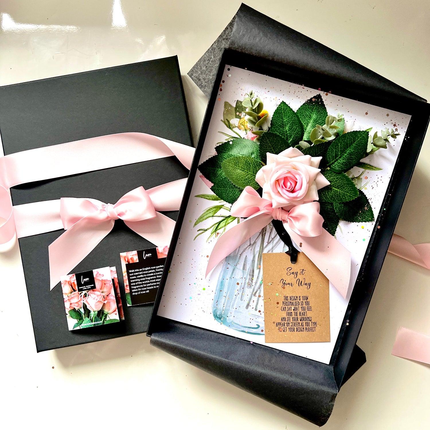 A floral gift box set featuring a black presentation box with a light pink ribbon and matching floral card, alongside an open box displaying a pink rose, green leaves, a small blue glass jar, and a kraft gift tag on a white background.