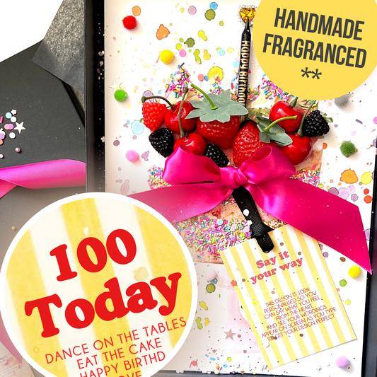 A close-up of a handmadeBirthday Cake Card Scented with Cake Mixture — for a Special 100th Birthday with a bright pink ribbon, topped with strawberries, cherries, and blackberries, alongside a a yellow striped note card.