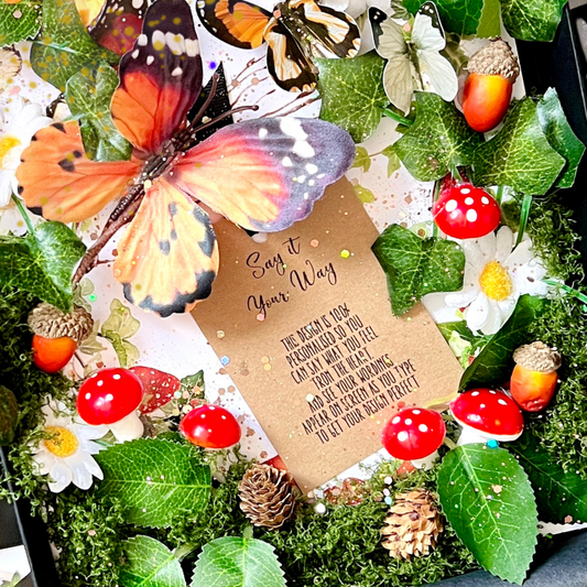 Close up of the magical enchanted luxury handmade personalised card design. A special design surrounded by artificial moss, green leaves, white daisies, red-and-white mushrooms, acorns, and large orange-and-black butterflies for something truly unique with the luxury touch