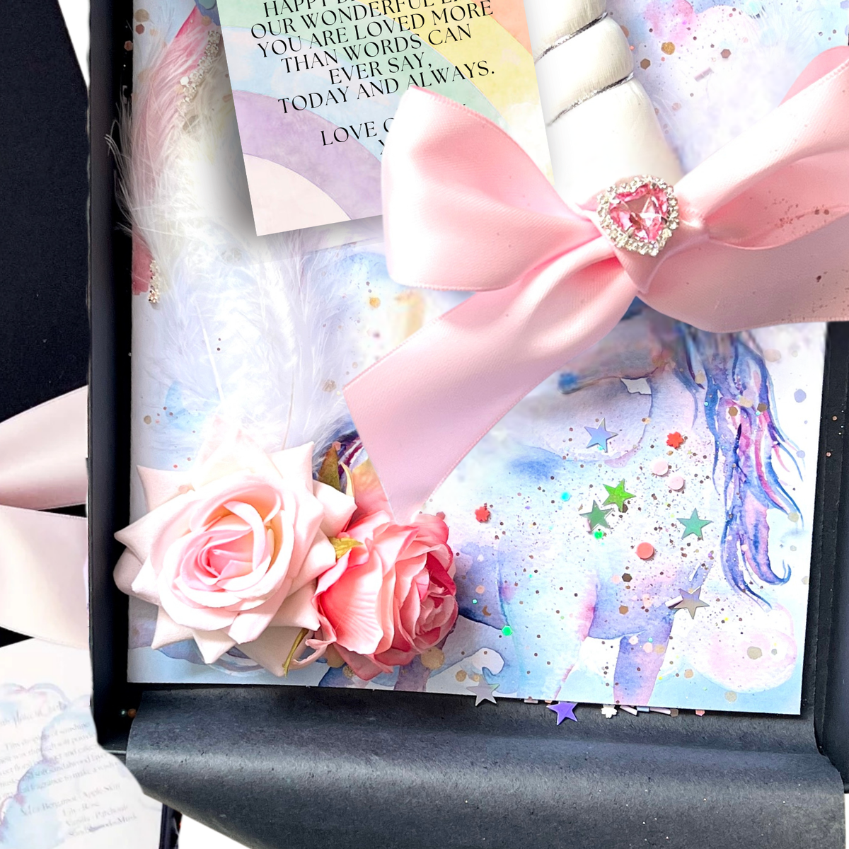A close-up of a decorative gift box that the magical unicorn card sits in featuring pastel unicorn artwork, a large pink satin bow with a jeweled center, pink fabric roses, white feathers, and a rainbow printed message card.