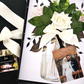 Muslim Wedding Card with Luxury Scented Floral arrangement with white roses and green leaves in a clear vase, surrounded by wedding-themed cards and photos.