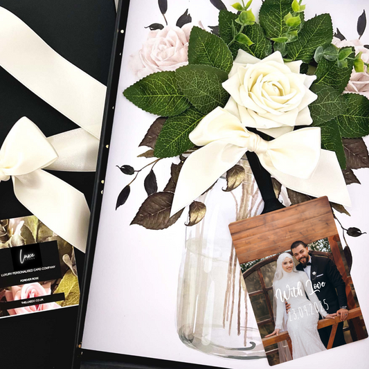 Muslim Wedding Card with Luxury Scented Floral arrangement with white roses and green leaves in a clear vase, surrounded by wedding-themed cards and photos.