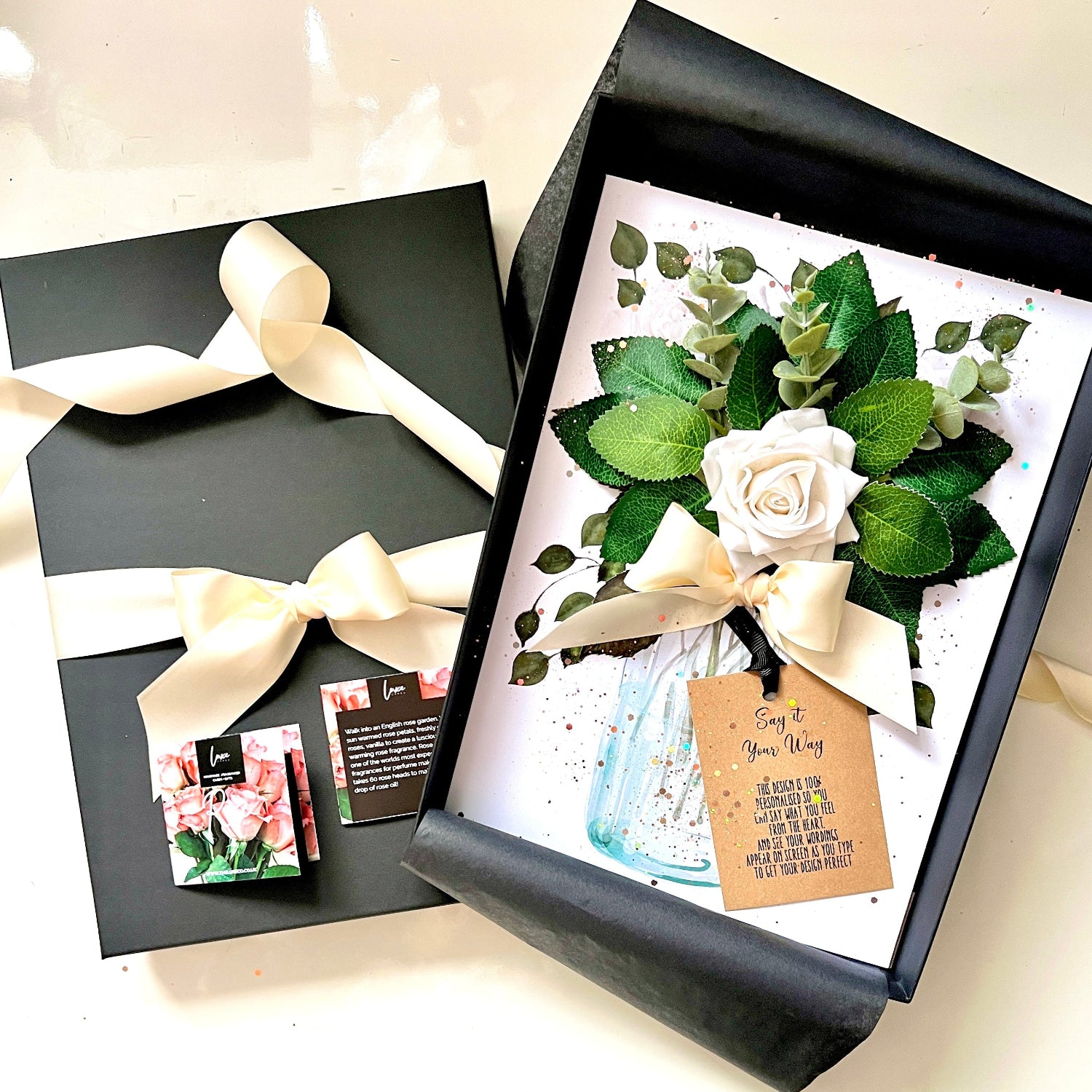 A floral gift box set on a light beige surface, featuring a black gift box with a cream ribbon and a matching small card, alongside an open black box displaying a white card with a faux rose, green leaves, a small blue glass jar, and a kraft gift tag.
