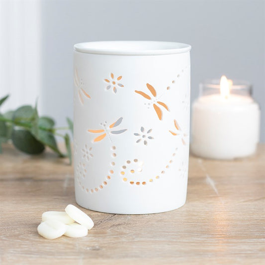 White ceramic dragonfly oil burner with floral cutout design on a wooden surface with a lit candle in the background.