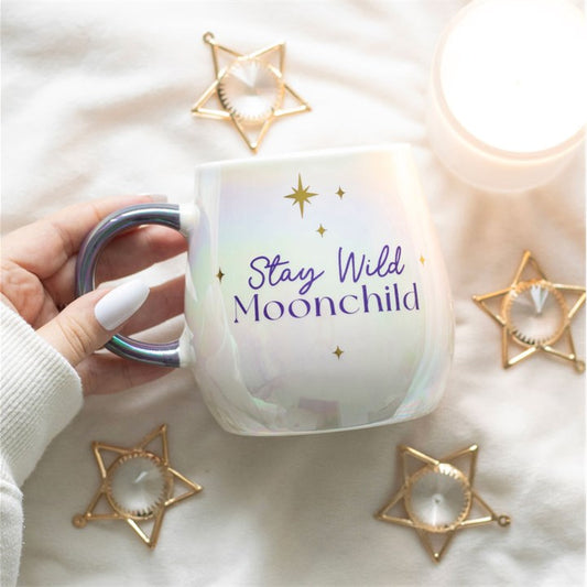 Mug with 'Stay Wild Moonchild' text held by a hand, surrounded by decorative star-shaped objects on a soft surface.
