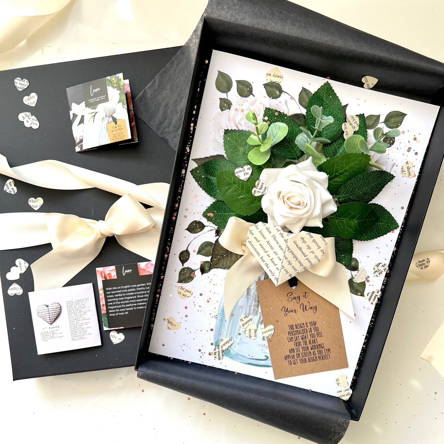 A square floral gift box set displayed on a light surface, featuring a black box with tissue paper, a white rose bouquet with green leaves and a cream ribbon, and several printed cards and labels.