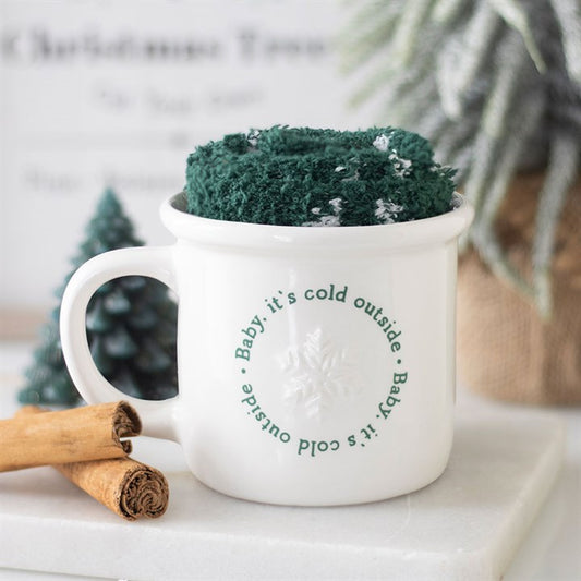White mug with text and green knitted cozy on a white surface with cinnamon sticks and a plant.