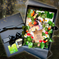An enchanted magical woodland butterfly handmade luxury personalised greetings card for birthdays, mothers day and more. Each black gift box contains a decorative arrangement of artificial green leaves, white daisies, red toadstools, pinecones, and several orange-and-black butterflies on a card. With a centre piece printed silk orange butterfly. A matching black ribboned box sits beside it on a dark textured background. Fragranced with Woodland. For a luxury card to wow the recipient the moment they open it