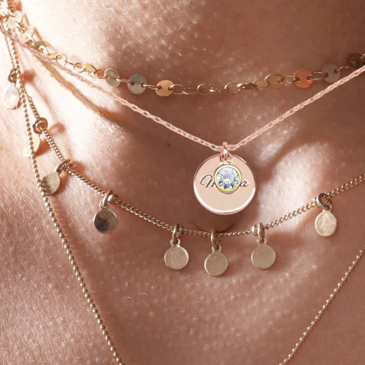 Close-up of a person wearing multiple gold necklaces with our personalised name or couples initials engagement Necklace circular pendants on a soft skin background.