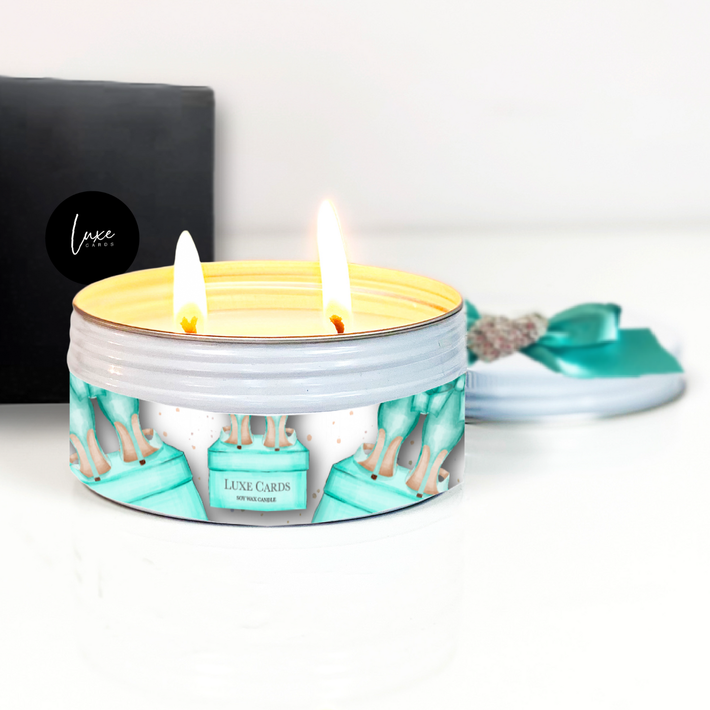 Lit candle in a decorative tin with 'Luxe Cards' branding on a white background