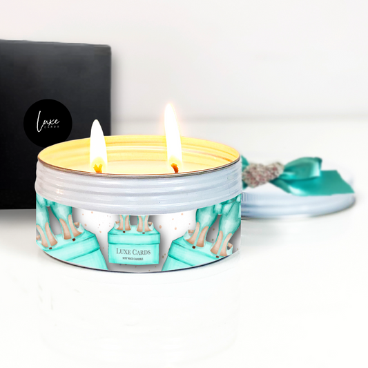Lit candle in a decorative tin with 'Luxe Cards' branding on a white background