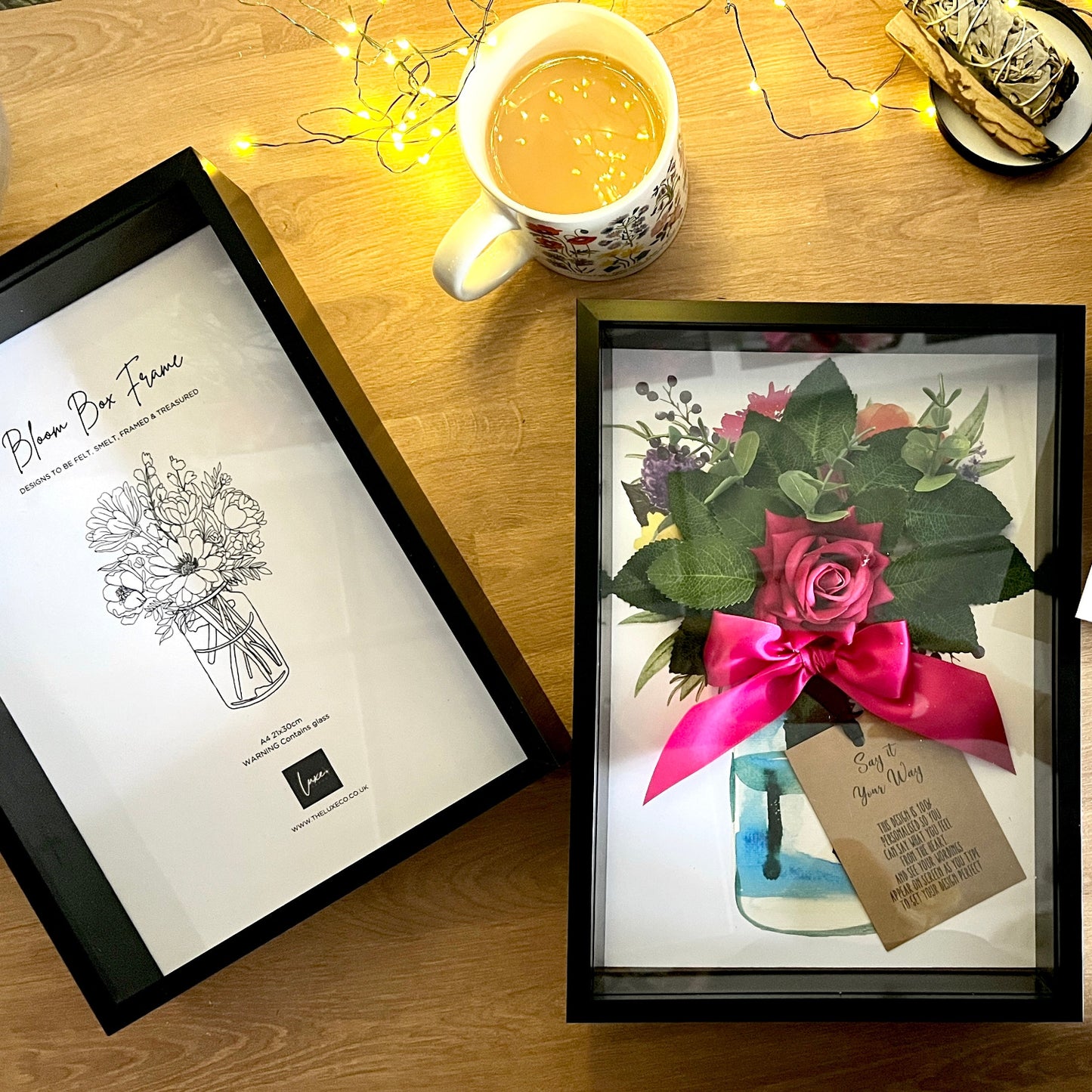 A framed floral gift box displayed on a wooden table, surrounded by a mug of tea, fairy lights, and another floral print. The central product is a black-framed box containing a small bouquet with a bright pink ribbon and a hanging tag.